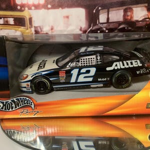 May include: A black and white Hot Wheels die-cast car with the number 12 and the words "Alltel" and "Mobil 1" on the side. The car is in a clear plastic display case with a yellow and orange background. The car is a 2002 NASCAR race car.