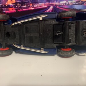 May include: A blue toy car with black tires and red rims, viewed from underneath. The car has silver trim and is set against a blurred background. The toy car is a classic vehicle.