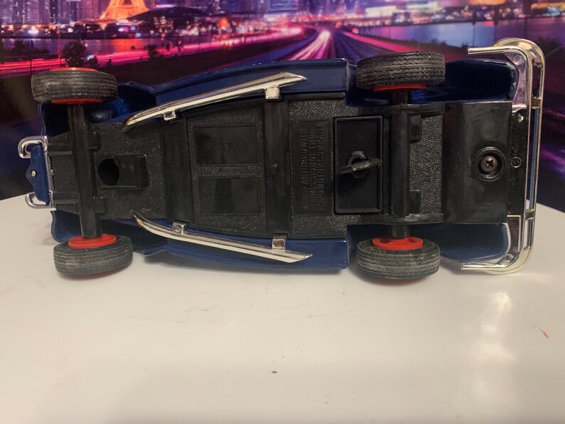 May include: A blue toy car with black tires and red rims, viewed from underneath. The car has silver trim and is set against a blurred background. The toy car is a classic vehicle.