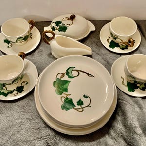May include: A vintage cream-colored dinnerware set featuring a green ivy leaf design. The set includes cups, saucers, plates, a gravy boat, and a covered dish. The cups have gold-colored handles.