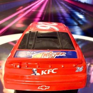 May include: A red toy race car with the number 26 on top and the rear. The car features KFC and Twister branding. The Chevrolet logo is visible on the rear. The car is a miniature replica.