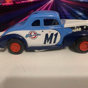 May include: A vintage-style toy car in blue, white, and navy. The car features red wheels and the number "M1" on the side. The car has a "Zip plus Mile Ale" logo and the word "Fireball" on the roof.