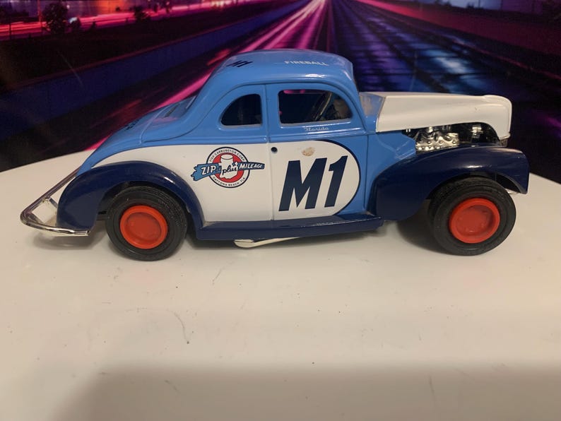 May include: A vintage-style toy car in blue, white, and navy. The car features red wheels and the number "M1" on the side. The car has a "Zip plus Mile Ale" logo and the word "Fireball" on the roof.