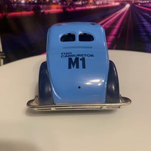 May include: A light blue toy car with the text "FISH CARBURETOR" and "M1" printed on the back. The car has a silver bumper and dark blue fenders. The car is a vintage style toy.