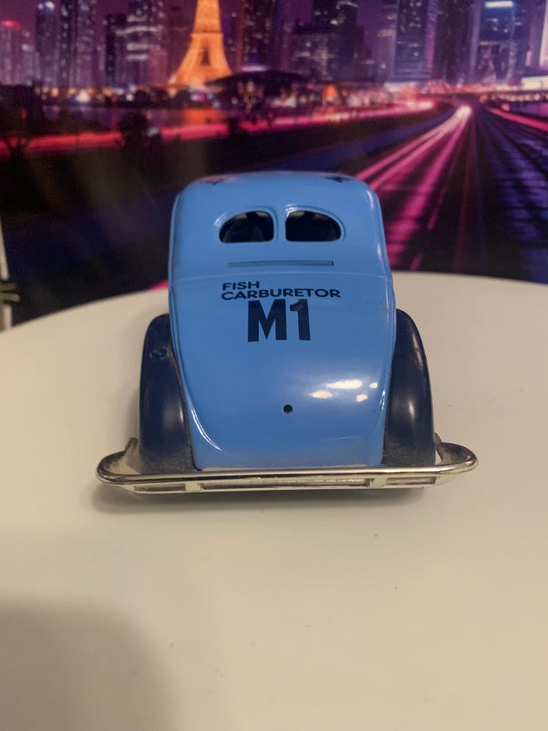 May include: A light blue toy car with the text "FISH CARBURETOR" and "M1" printed on the back. The car has a silver bumper and dark blue fenders. The car is a vintage style toy.