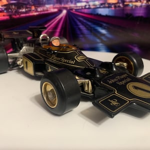 May include: A black and gold Lotus Formula 1 race car model. The car features the "John Player Special" logo and the number "1". The wheels are black with gold rims. A small figure is visible in the cockpit.