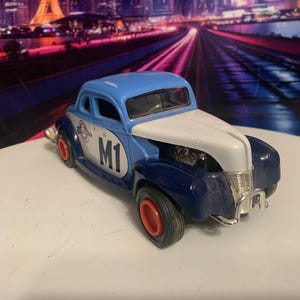 May include: A blue, white, and navy blue vintage-style toy car with red wheels. The car has the number "M1" on the side and a logo. The background features a blurred cityscape.