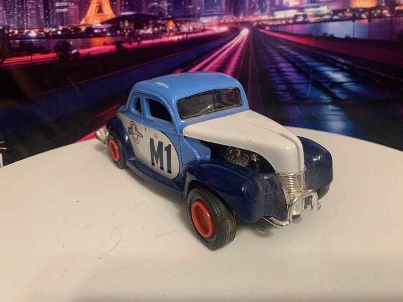 May include: A blue, white, and navy blue vintage-style toy car with red wheels. The car has the number "M1" on the side and a logo. The background features a blurred cityscape.