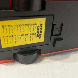 May include: The underside of a red and black toy vehicle featuring a yellow rectangular label printed with the Morse Code alphabet. The label displays letters A through Z alongside their corresponding dot and dash sequences. A black switch is visible.