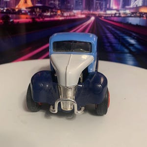 May include: A vintage-style toy car in blue, white, and navy. The car has a silver grill and red wheels. The toy car is a classic model, likely from the mid-20th century.