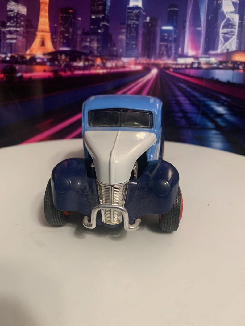 May include: A vintage-style toy car in blue, white, and navy. The car has a silver grill and red wheels. The toy car is a classic model, likely from the mid-20th century.