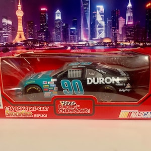 May include: A 1:24 scale die-cast replica of a NASCAR stock car. The car is black and teal with the number 90 on the side. The box is red and features the Racing Champions and NASCAR logos.