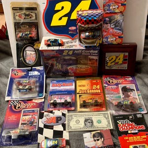 May include: A collection of Jeff Gordon NASCAR memorabilia, including die-cast cars, a signed Route 24 sign, a tin box, and a money clip. The items are displayed on a table with a dollar bill and a card.