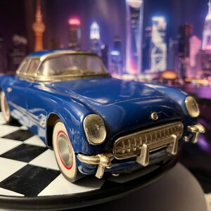 May include: A blue die-cast model car featuring a silver front grille, chrome-colored bumpers, and white-walled tires with red accents. The vehicle has a hardtop roof and clear windows, showcasing a detailed exterior design with a glossy finish.