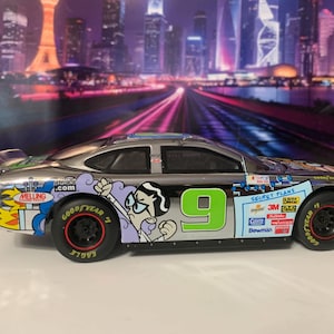 May include: A silver race car toy with the number 9 in green on the side. The car features cartoon graphics and sponsor logos. The tires are black with red rims and Goodyear branding.
