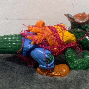 May include: A green alligator action figure wearing a brown cowboy hat and holding a red, orange, and yellow figure in its arms. The figures are tangled in pink string.