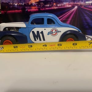 May include: A blue, white, and navy toy car with the number "M1" on the side. The car has red wheels and measures approximately 7 inches in length. The car has the words "Zip Italian Mille Miglia" on the side.
