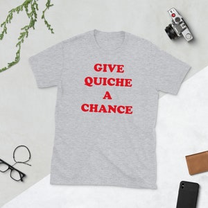 Puede incluir: Una camiseta gris con el texto "GIVE QUICHE A CHANCE" impreso en rojo.