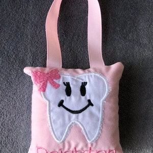 May include: Pink tooth-shaped pillow with a smiling face, a pink bow, and the name "Delighton" embroidered on the front. The pillow has a pink ribbon loop for hanging.