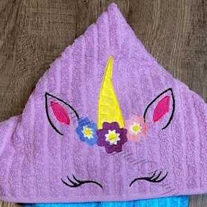 Puede incluir: Toalla con capucha morada con un unicornio bordado en la parte delantera. El unicornio tiene un cuerno amarillo, orejas rosas y una corona de flores con flores moradas, amarillas y azules.