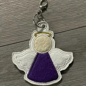 May include: A white and purple angel-shaped keychain with a gold halo and silver clasp. The angel has white wings, a beige head, and a purple dress. The keychain is made of felt and has a stitched outline.