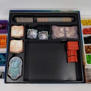Puede incluir: Un organizador de juegos con m&uacute;ltiples compartimentos para guardar las piezas y las cartas del juego. El organizador tiene una bandeja negra con un borde gris y una bandeja blanca con m&uacute;ltiples compartimentos. El organizador est&aacute; lleno de diferentes piezas de juego de colores, incluyendo rojo, naranja, azul, verde y marr&oacute;n.