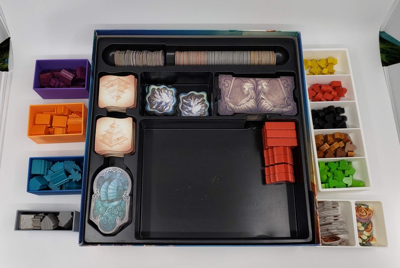 Puede incluir: Un organizador de juegos con m&uacute;ltiples compartimentos para guardar las piezas y las cartas del juego. El organizador tiene una bandeja negra con un borde gris y una bandeja blanca con m&uacute;ltiples compartimentos. El organizador est&aacute; lleno de diferentes piezas de juego de colores, incluyendo rojo, naranja, azul, verde y marr&oacute;n.