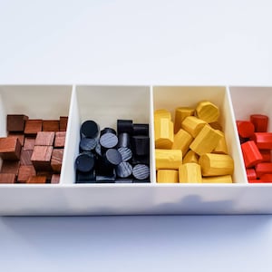 May include: A white tray with four compartments, each filled with wooden game pieces. The compartments contain brown, black, yellow, and red pieces.