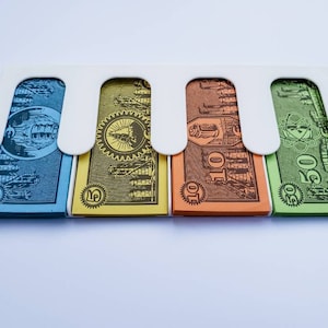 May include: Four Monopoly game money bills in a white plastic holder. The bills are blue, yellow, orange, and green. The bills are labelled with the amounts &pound;1, &pound;5, &pound;10, and &pound;50.