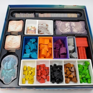 Puede incluir: Un inserto organizador de juego con compartimentos para guardar las piezas y las cartas del juego. El inserto tiene compartimentos para piezas de madera azules, naranjas, moradas, grises y rojas, as&iacute; como compartimentos para cartas con ilustraciones de edificios y personas.