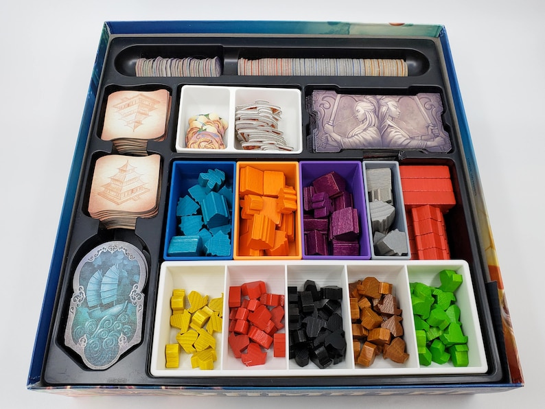 Puede incluir: Un inserto organizador de juego con compartimentos para guardar las piezas y las cartas del juego. El inserto tiene compartimentos para piezas de madera azules, naranjas, moradas, grises y rojas, as&iacute; como compartimentos para cartas con ilustraciones de edificios y personas.
