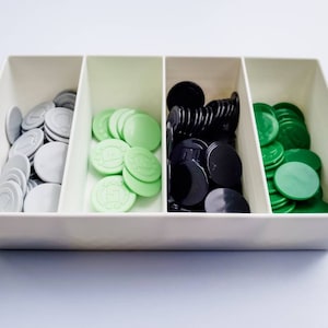 May include: A white plastic tray with three compartments, each filled with a different colour of game chips. The left compartment contains silver chips, the middle compartment contains light green chips, and the right compartment contains black chips.