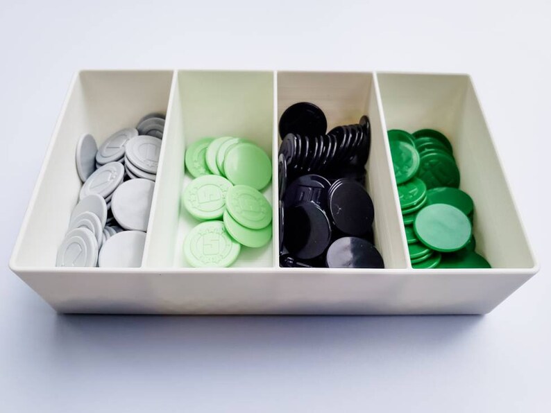 May include: A white plastic tray with three compartments, each filled with a different colour of game chips. The left compartment contains silver chips, the middle compartment contains light green chips, and the right compartment contains black chips.