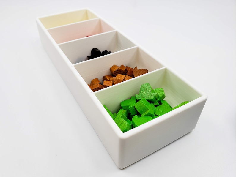 Puede incluir: Una caja rectangular blanca con cinco compartimentos. Los compartimentos est&aacute;n llenos de peque&ntilde;as piezas de madera de diferentes colores: verde, marr&oacute;n, negro, rojo y blanco.