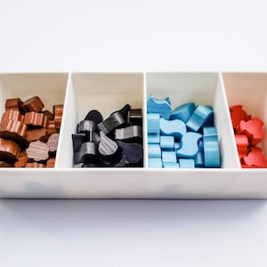 May include: A white tray with four compartments, each filled with wooden game pieces. The compartments contain brown, black, blue, and red pieces.