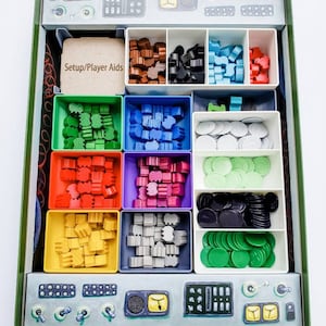 May include: A green box with a compartmentalised insert for storing game pieces. The compartments are filled with various coloured plastic pieces, including red, green, blue, yellow, and grey. The box also has a label that says "Setup/Player Aids".