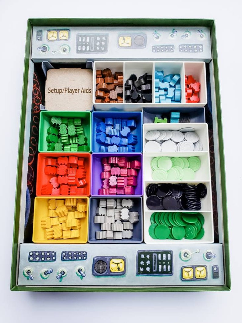 May include: A green box with a compartmentalised insert for storing game pieces. The compartments are filled with various coloured plastic pieces, including red, green, blue, yellow, and grey. The box also has a label that says "Setup/Player Aids".