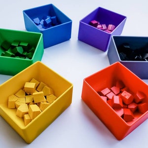 May include: Six small square containers, each filled with colourful wooden blocks. The containers are arranged in a circle on a white surface. The colours of the containers are blue, green, purple, grey, yellow, and red.