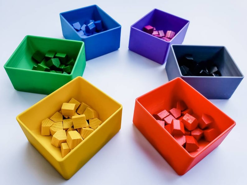 May include: Six small square containers, each filled with colourful wooden blocks. The containers are arranged in a circle on a white surface. The colours of the containers are blue, green, purple, grey, yellow, and red.