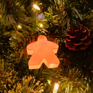 May include: A glowing orange game piece ornament in the shape of a person hangs on a Christmas tree. A brown pine cone and twinkling lights are also visible among the green pine needles.