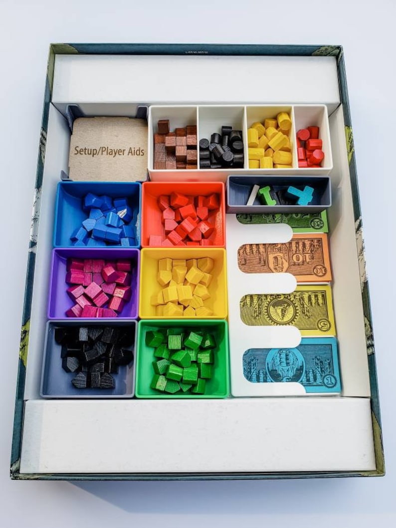 May include: A game box organiser with compartments for storing game pieces. The organiser has compartments for blue, red, green, purple, black, and brown pieces. There are also compartments for money and player aids. The text "Setup/Player Aids" is printed on a brown card.