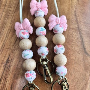 May include: Three beaded lanyards with pink bow accents. The lanyards feature wooden beads and floral-patterned beads. Each lanyard has a bronze-colored clasp. The background is a wooden surface.