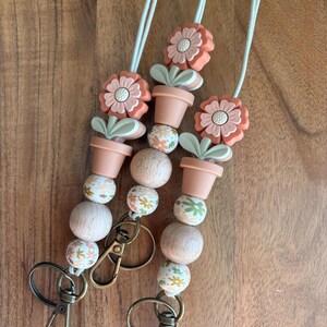 May include: Three beaded lanyards with a floral theme. Each lanyard features peach-colored flower and pot charms, wooden beads, and decorative beads with floral patterns. The lanyards have bronze-colored clasps and white cords.