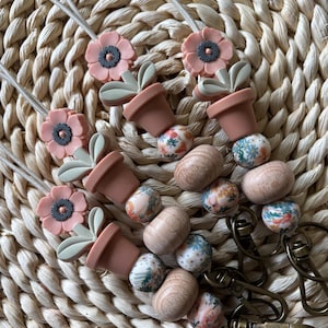 May include: Several handmade lanyards with floral and plant-themed charms. Each lanyard features a pink flower, a miniature terracotta pot with green leaves, and wooden and patterned beads. The lanyards are attached to bronze-colored clasps.