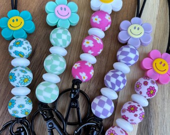 Puede incluir: Una colección de coloridas correas para el cuello con dijes de flores y caras sonrientes. Cada correa para el cuello presenta un cordón negro, un cierre y cuentas decorativas en varios patrones y colores, incluidos diseños de cuadros y florales.