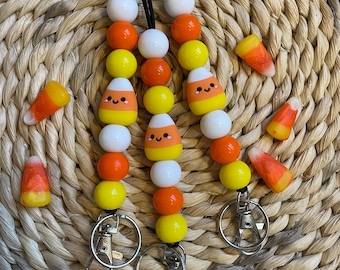 May include: Three candy corn keychains with black cords and silver clasps. Each keychain has white, orange, and yellow beads with a candy corn charm with a smiling face.