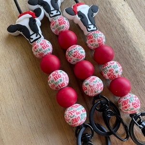 May include: Three Christmas-themed lanyards with black cords. Each lanyard features a cow head wearing a Santa hat, red and white patterned beads with "HO HO HO" text, and solid red beads. The lanyards have black metal clasps.