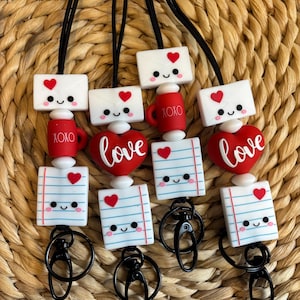 May include: A collection of Valentine's Day-themed lanyards. Each lanyard features a black cord with decorative beads. The beads include white envelopes with hearts, red mugs with "XOXO", red hearts with "love", and lined paper with smiling faces.