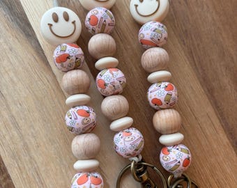 Puede incluir: Tres cordones con cuentas con temática escolar. Cada cordón presenta una cuenta blanca con una carita sonriente, cuentas de madera y cuentas rosas con diseños de manzanas y lápices. Los cordones tienen cierres de color bronce.