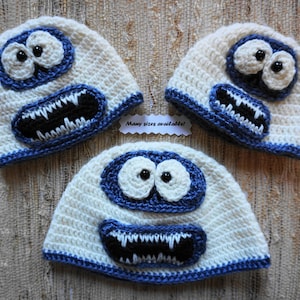May include: Three white crocheted hats with blue and black details. The hats feature a cartoon monster face with large eyes, a toothy grin, and a blue nose. The text "Many sizes available!" is visible in the image.